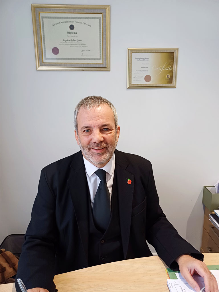 Steve Jones is sitting at his desk with his two Diplomas from the National Association of Funeral Directors framed behind him on the wall.